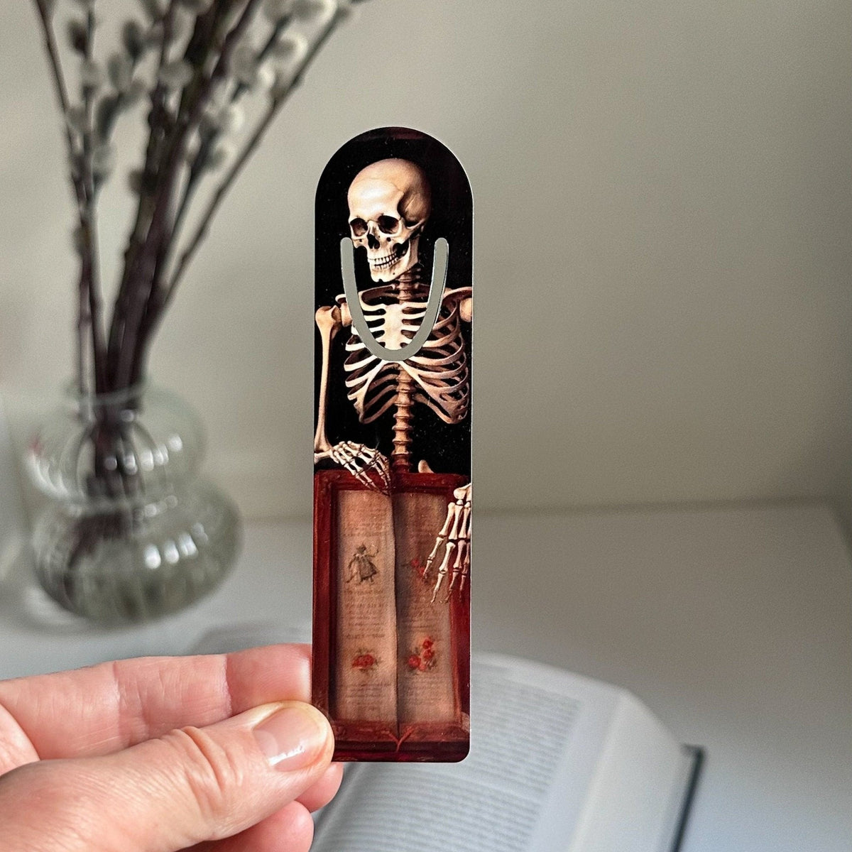 Skeleton with the Book metal bookmark held in a hand above an open book