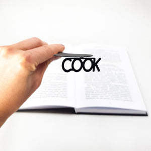 Hand holding a black metal spoon-shaped bookmark with the word “COOK” cut out, with an open book blurred in the background.
