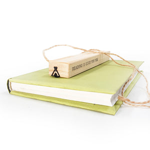 Black metal teepee bookmark resting on the spine of a green book, with a wooden packaging block labeled “Reading is good for you” and twine string beside it.i