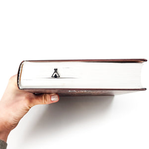 Hand holding a closed hardcover book with a black metal teepee bookmark visible between the pages on the book’s spine.
