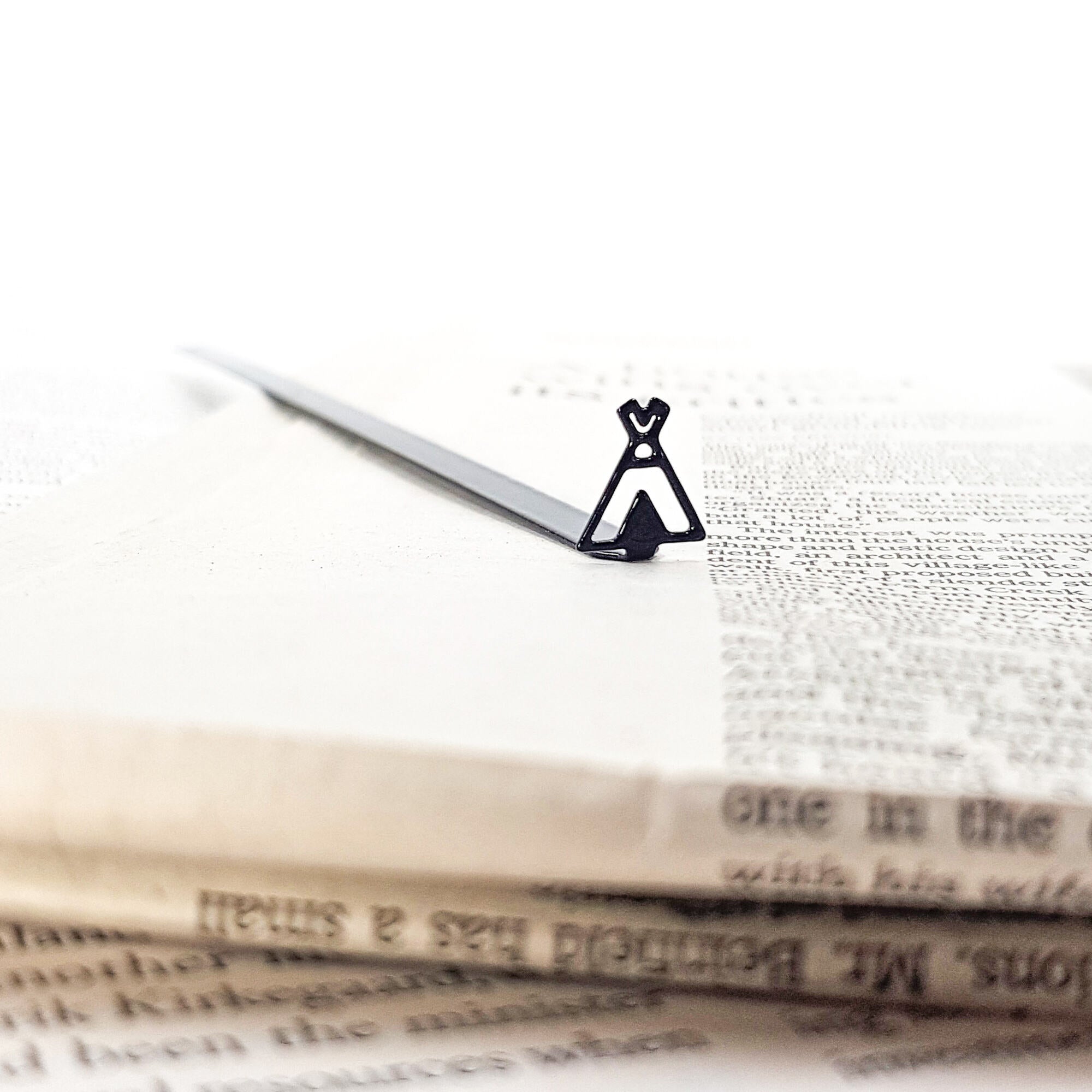 Close-up of a black teepee-shaped metal bookmark lying on an open book page, with stacked vintage-style papers in the foreground.