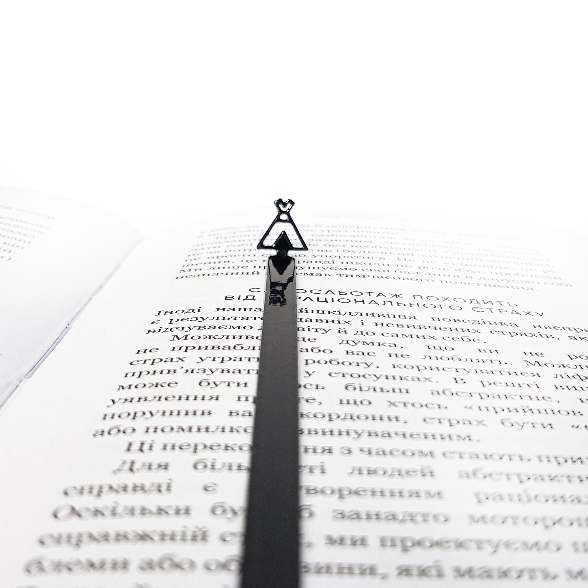 black teepee-shaped metal bookmark lying across an open book with Cyrillic text, tip pointing toward the top of the page.