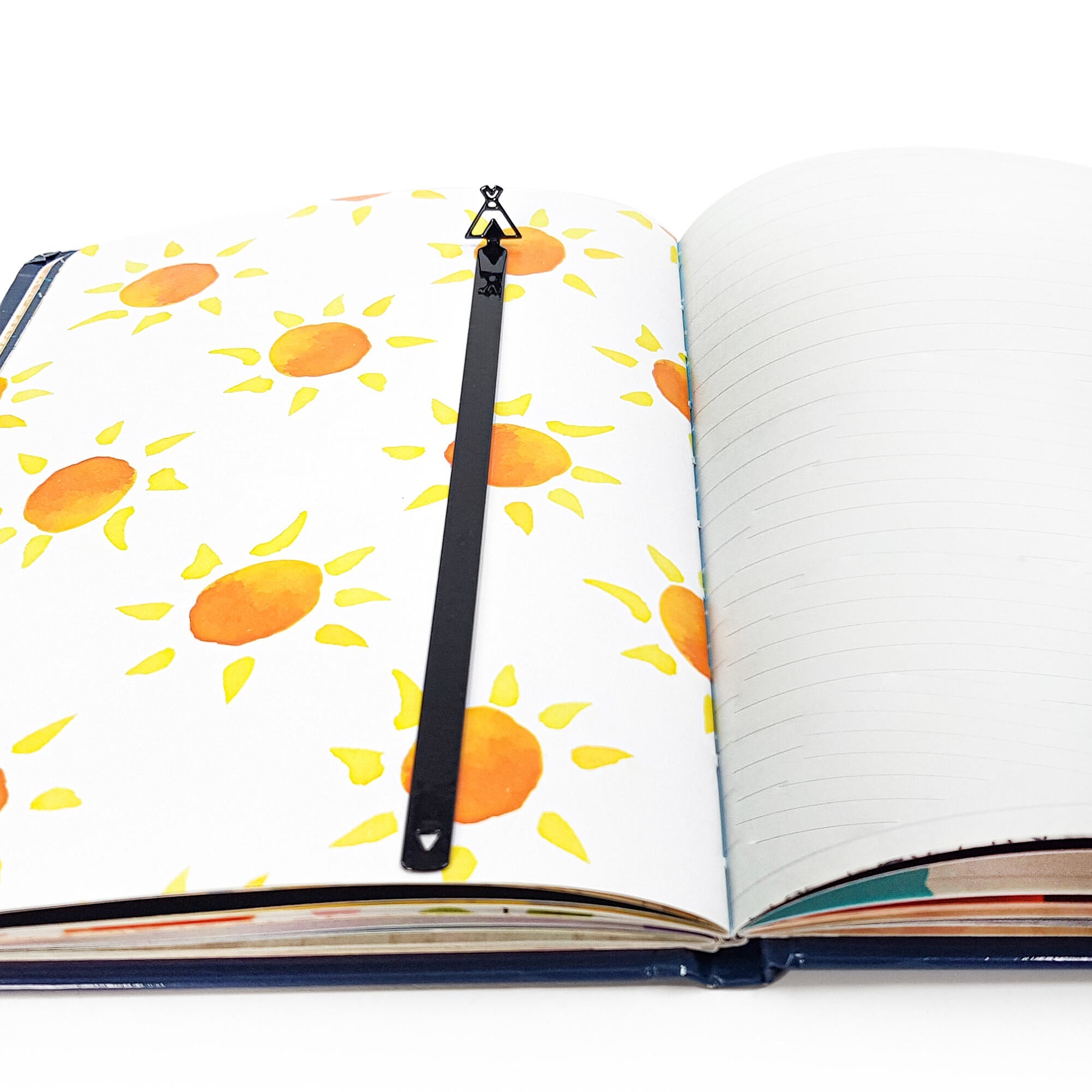 Teepee-shaped black metal bookmark placed along the center fold of an open notebook with sun illustrations on one page and lines on the other.