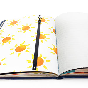 Teepee-shaped black metal bookmark placed along the center fold of an open notebook with sun illustrations on one page and lines on the other.