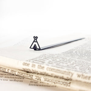 Black metal teepee-shaped bookmark resting on an open book, with vintage newspaper text in the foreground and a minimal white background.
