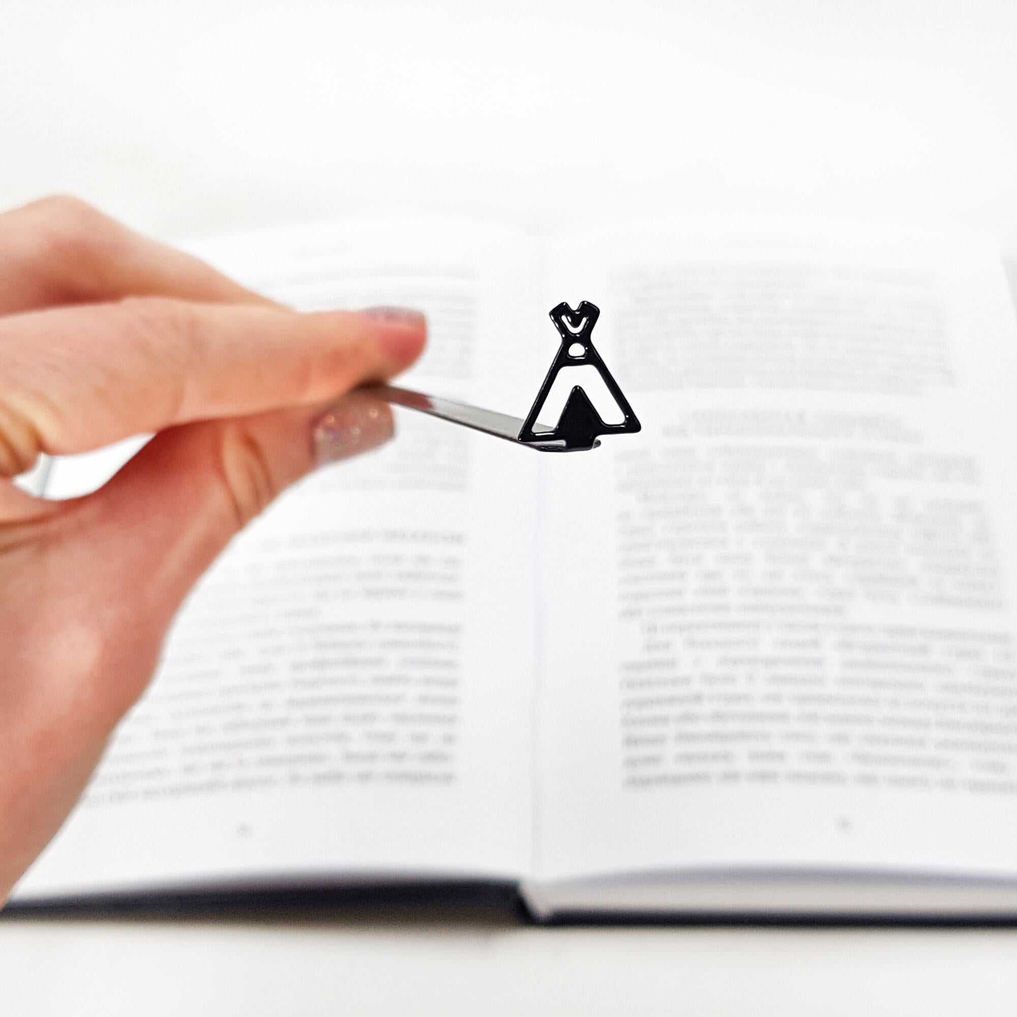 Hand holding a black teepee-shaped metal bookmark above the center of an open book with blurred text.