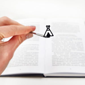 Hand holding a black teepee-shaped metal bookmark above the center of an open book with blurred text.