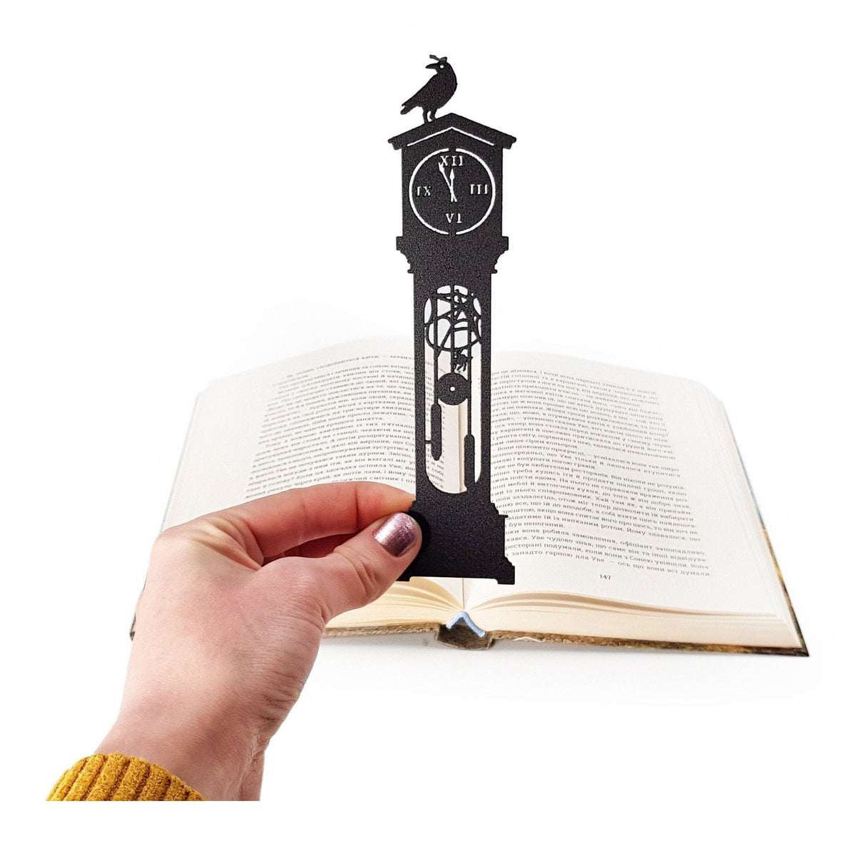 The Raven's Pendulum Dance metal bookmark held in a hand above an open book