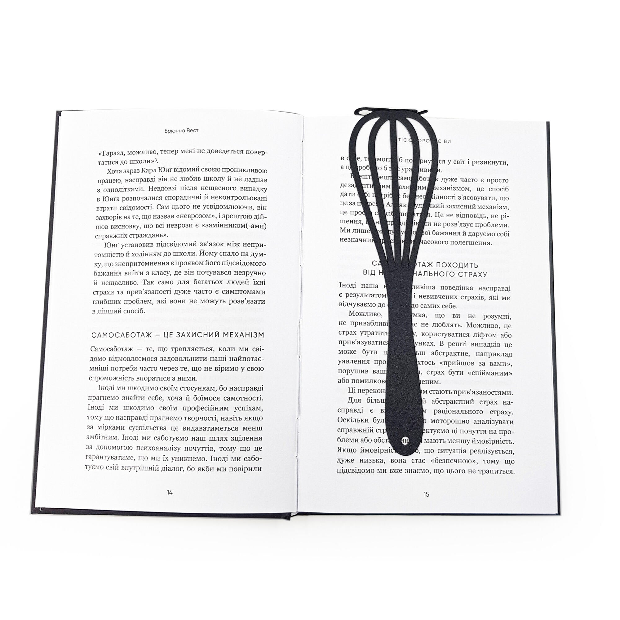 A black metal whisk-shaped bookmark lies flat across the center of an open book with text in Ukrainian. Designed and made in Ukraine. 