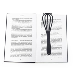 A black metal whisk-shaped bookmark lies flat across the center of an open book with text in Ukrainian. Designed and made in Ukraine. 