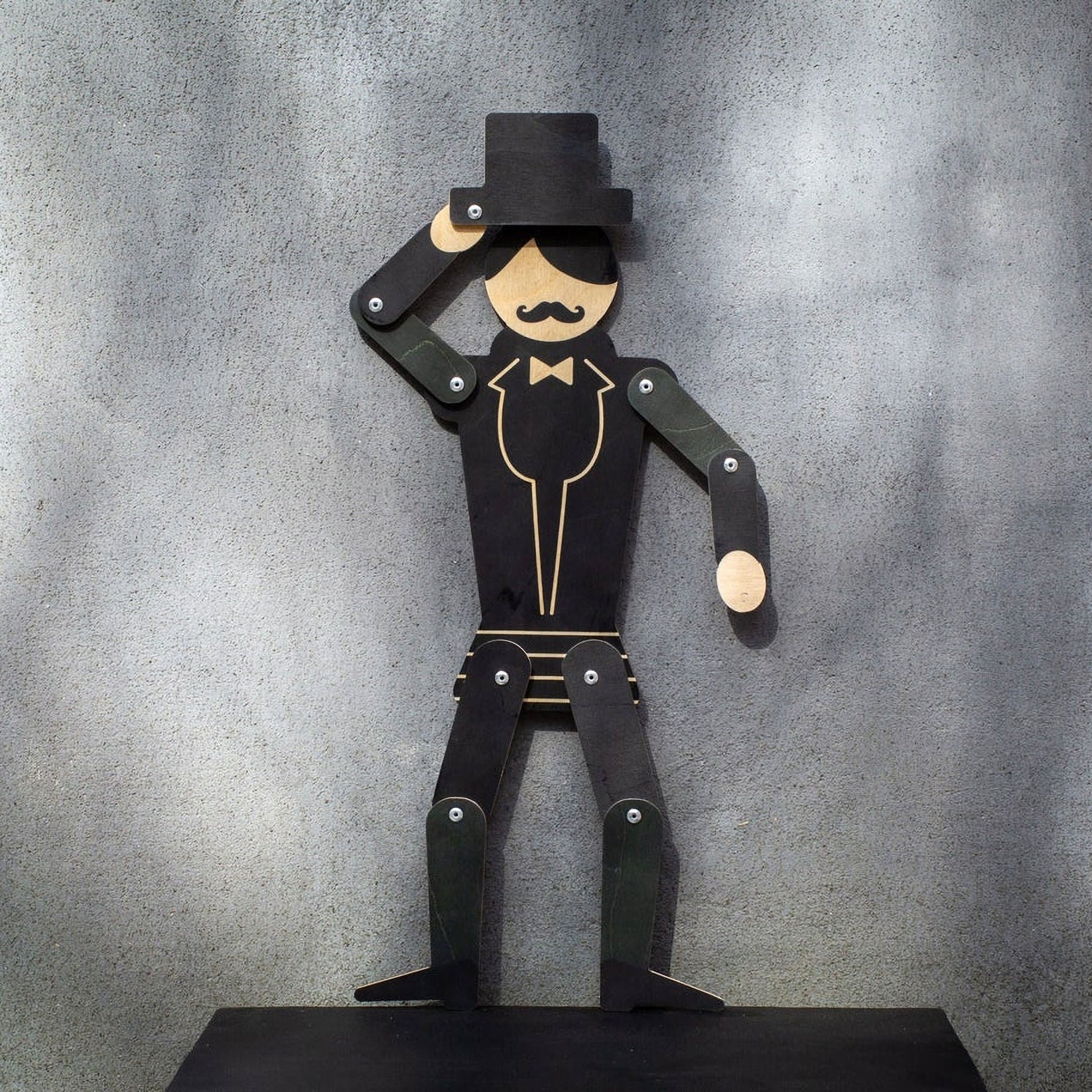 Wooden gentleman puppet on dark shelf against grey wall, tipping top hat with all joints visible