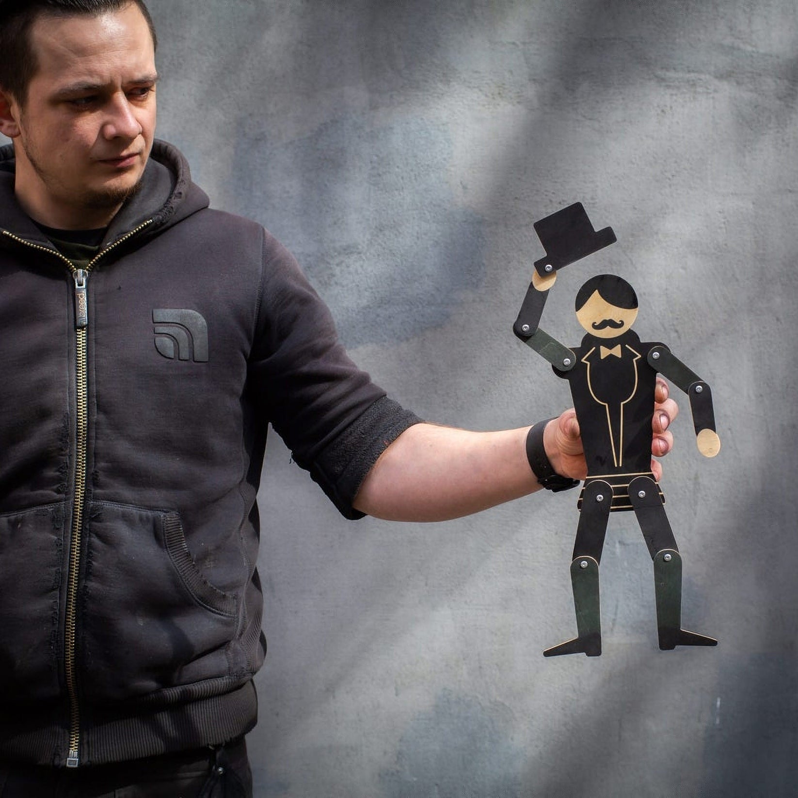 Maker holding wooden gentleman puppet at arm's length showing scale — tuxedo figure tipping top hat