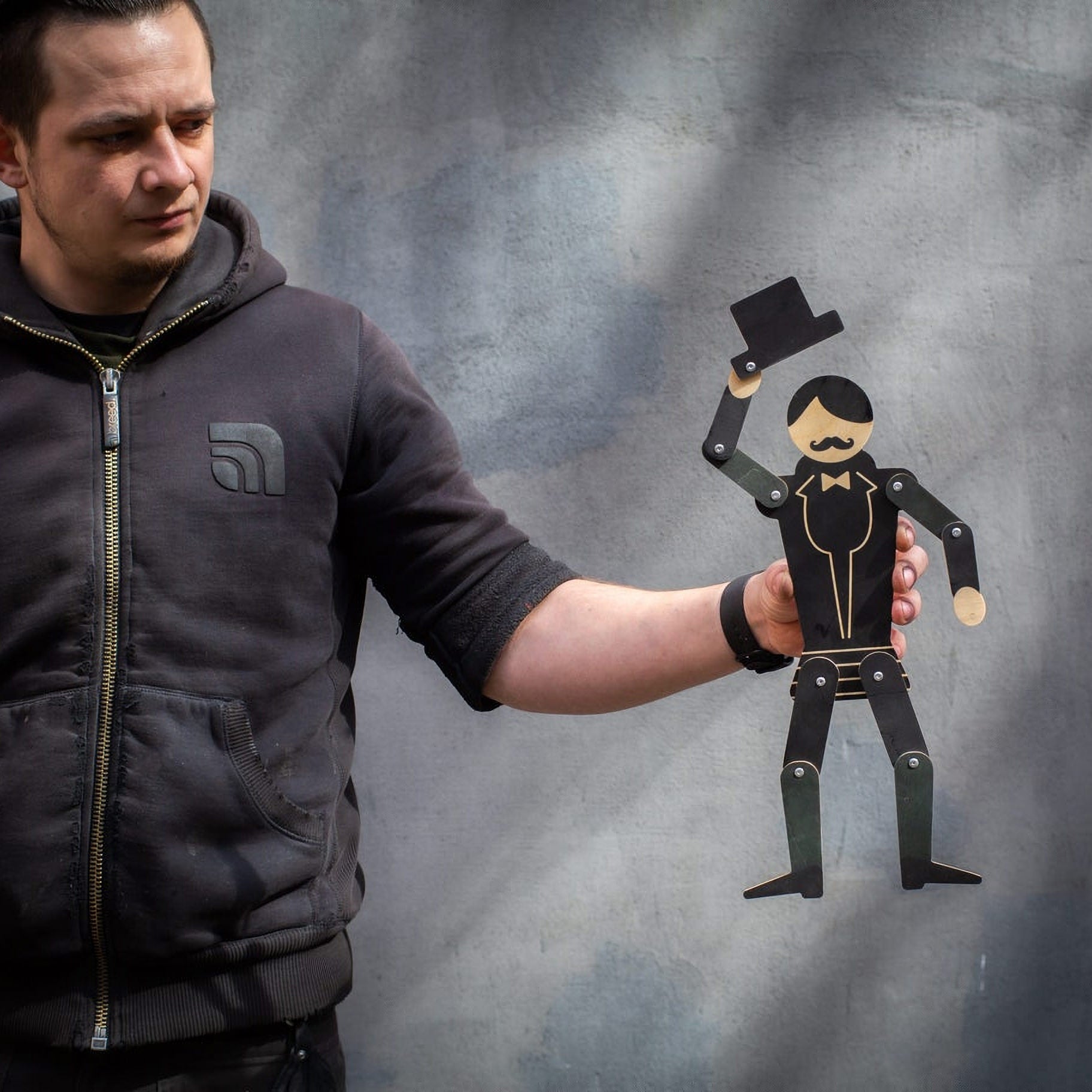 Maker holding wooden gentleman puppet at arm's length showing scale — tuxedo figure tipping top hat